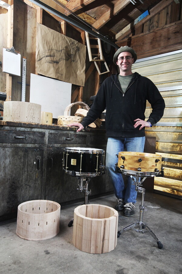 HIS CREATIONS WILL SNARE YOUR ATTENTION. Eric Lee stands in his workshop with examples of a multi-vented stave shell (lower left), a finished “Joey G Signature Series” (in black), and a segmented snare (right).